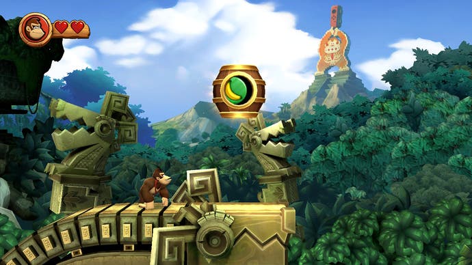 Donkey Kong looks up at a large barrel in mid air in a jungle level, while a NES statue of himself holding a Switch Joy-Con looms in the background in Donkey Kong Country Returns HD.