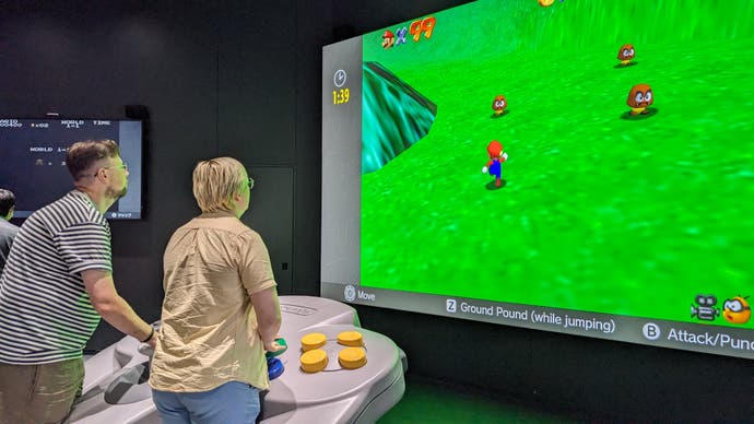 Two people play Super Mario 64 with a giant N64 controller at the Nintendo Museum.